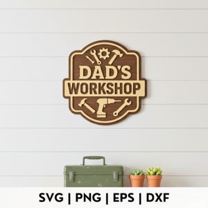 May include: A wooden sign with the text "DAD'S WORKSHOP" and various tool illustrations. The sign is brown and tan, with a layered design. Below the sign is a green toolbox and two small potted plants.