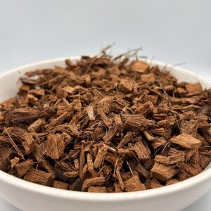 May include: A white ceramic bowl filled with brown coconut husk chips. The chips are various sizes and shapes, with a rough, textured appearance. The bowl is set against a plain white background, highlighting the natural color and texture of the coconut husk.