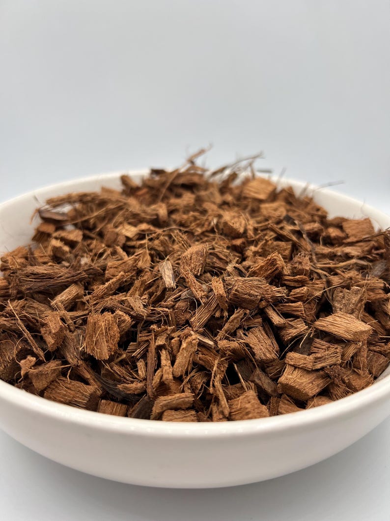 May include: A white ceramic bowl filled with brown coconut husk chips. The chips are various sizes and shapes, with a rough, textured appearance. The bowl is set against a plain white background, highlighting the natural color and texture of the coconut husk.