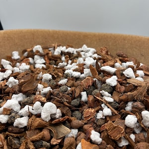 May include: Close-up of a mix of potting soil components, including white perlite, brown bark pieces, and small, dark pebbles. The blend is in a brown container, ideal for plants needing well-draining soil. The texture is coarse and varied.
