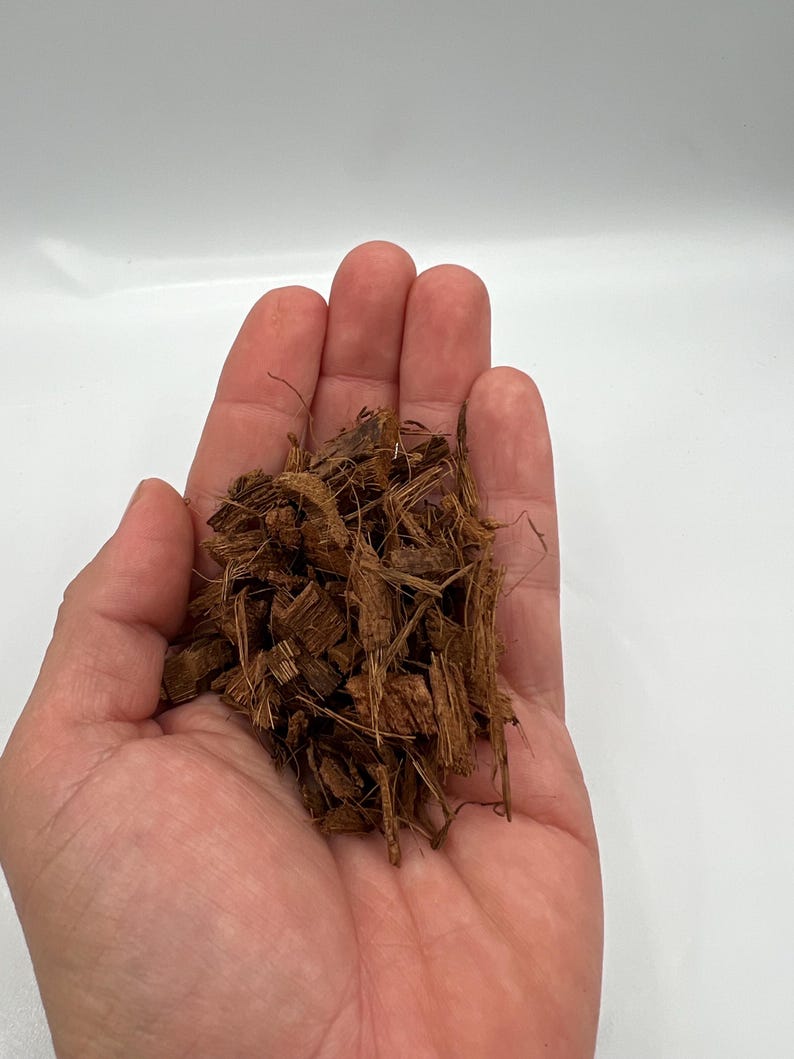 May include: A handful of brown, shredded coconut husk, with a light brown color and a rough texture. The coconut husk is held in an open hand against a white background. The husk appears to be for gardening or pet bedding.
