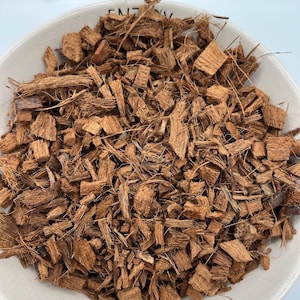 May include: A white ceramic bowl filled with brown coconut coir. The coir is composed of small, irregularly shaped pieces of coconut husk and fibers. The bowl is set against a white background.