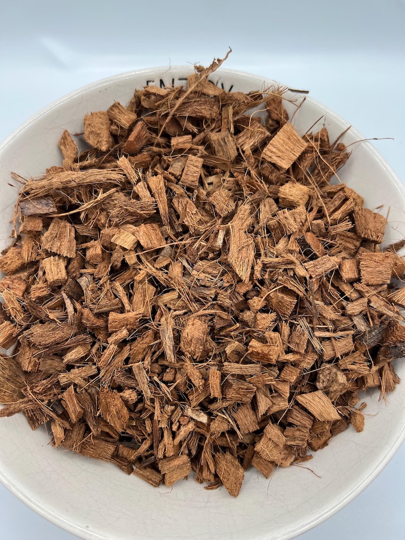 May include: A white ceramic bowl filled with brown coconut coir. The coir is composed of small, irregularly shaped pieces of coconut husk and fibers. The bowl is set against a white background.