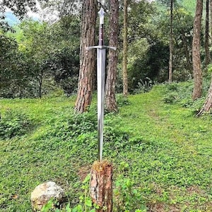 May include: A long, double-edged steel sword with a pointed tip, a straight crossguard, and a dark brown cylindrical handle topped with a silver-coloured pommel, standing vertically embedded into the top of a rough, textured wooden tree stump.