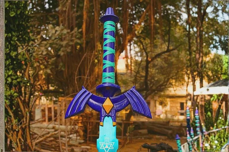 Pu&ograve; includere: Una spada giocattolo con elsa blu scuro e impugnatura avvolta in turchese brillante, una guardia blu scuro con un emblema centrale giallo a forma di diamante e una lama azzurra contrassegnata da un simbolo triangolare bianco vicino alla guardia.