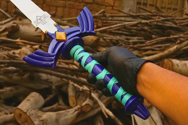 Pu&ograve; includere: Una replica di spada con lama argentata e emblema triangolare nero, una guardia blu scuro con un diamante dorato centrale e un'impugnatura avvolta da fasce alternate blu scuro e turchese chiaro, terminante con un pomolo blu scuro.