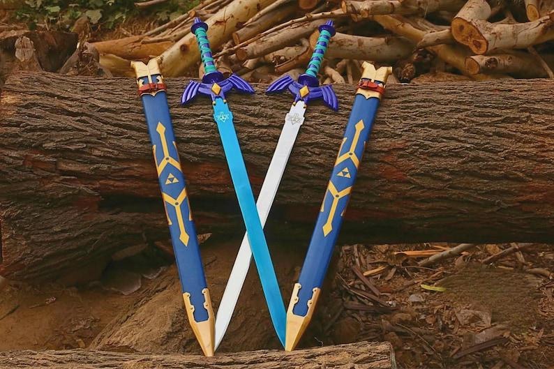 Pu&ograve; includere: Due spade con lame blu e argento, guardie viola e impugnature avvolte in verde acqua, ciascuna accompagnata da un fodero blu con accenti dorati e un emblema triangolare giallo, disposte in posizione incrociata su uno sfondo di legno.