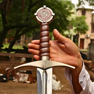 May include: A medieval-style sword with a silver blade and a brown leather-wrapped handle. The hilt features a decorative cross-shaped design with a circular emblem. The sword is held in front of a blurred outdoor background.