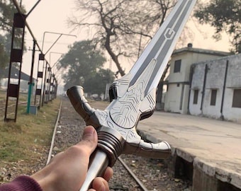 May include: A silver-colored sword with a long, pointed blade and intricate designs. The hilt features a crossguard and a textured grip. The blade has a central groove and decorative patterns. The sword is held against a backdrop of train tracks and a building.