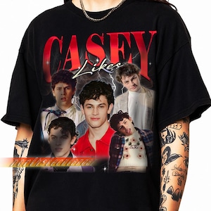 Può includere: T-shirt nero con grafica rossa "CASEY Likes" con effetto fulmine. Il design include più immagini di una persona. La maglietta è a girocollo.