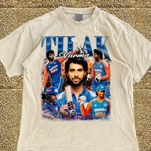 May include: Cream-colored t-shirt featuring a graphic of a cricket player with the name "Varma" above the image. The design includes images of the player in action, wearing blue and orange team uniforms. The word "THAK" is printed above the name.