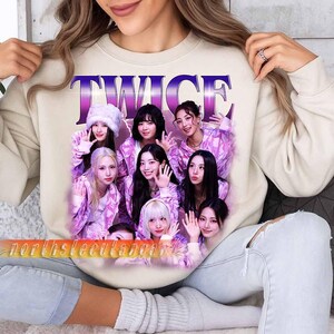 May include: Beige sweatshirt with a graphic of the K-pop group TWICE. The graphic shows the word "TWICE" in purple and pink, and images of the group members in pink and purple outfits. The sweatshirt is being worn by a person.