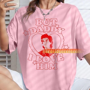 Może przedstawiać: Różowa koszulka z czerwonym napisem "BUT DADDY I LOVE HIM" i czerwonym wizerunkiem osoby w sercu. Koszulka ma swobodny styl.