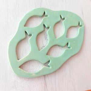 Puede incluir: Un salvamanteles de cerámica verde claro con un diseño único de peces. El salvamanteles presenta cortes en forma de peces, creando un patrón calado. Es de color verde pálido y se utiliza para colocar platos calientes.