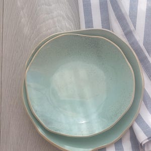 Op de afbeelding: Twee gestapelde keramische kommen met een organische, onregelmatige ronde vorm en een licht mintgroen gespikkeld glazuur. De kommen hebben een gladde binnenkant en een matte, lichtgekleurde rand, gepresenteerd naast een wit en blauw gestreepte stof.