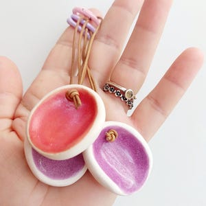 May include: Three oval ceramic pendants with white edges and glossy centers in shades of pink and purple. Each pendant features a small hole with a tan leather cord threaded through it, and the set is held together by small pastel plastic rings.