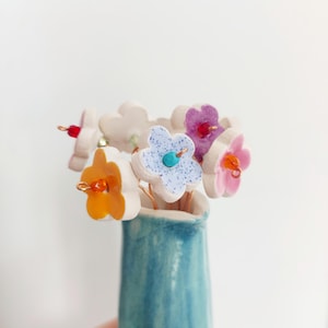 Könnte beinhalten: Eine kleine, blaue Keramikvase enthält einen Strauß handgefertigter Keramikblumen. Die Blumen sind weiß mit farbigen Zentren in Orange, Rosa, Lila und Blau. Jede Blume hat einen Drahtstiel und eine kleine Perle in der Mitte. Die Vase ist hellblau.