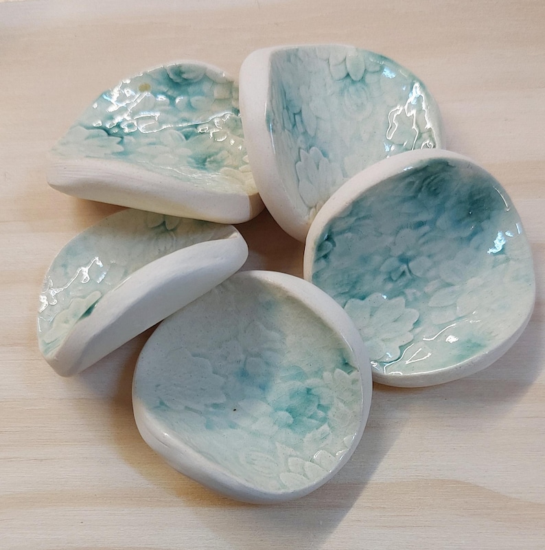 May include: Five small, handmade ceramic dishes. Each dish has a rounded shape with a white rim and a teal and white floral pattern inside. The dishes are arranged on a light-colored wooden surface.