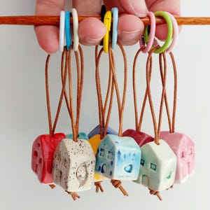 Könnte beinhalten: Eine Sammlung farbenfroher Keramik-Hausanhänger in verschiedenen Farben, darunter Rot, Beige, Blau und Rosa. Die Anhänger sind an Lederbändern befestigt und an farbigen Ringen angebracht.