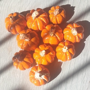 May include: A collection of small, orange ceramic pumpkins with textured surfaces and brown stems. The pumpkins are arranged on a light-colored surface, with shadows adding depth. Ideal for fall decor.