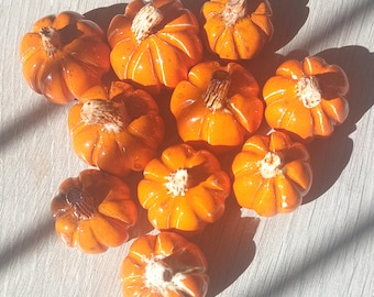 May include: A collection of small, orange ceramic pumpkins with textured surfaces and brown stems. The pumpkins are arranged on a light-colored surface, with shadows adding depth. Ideal for fall decor.