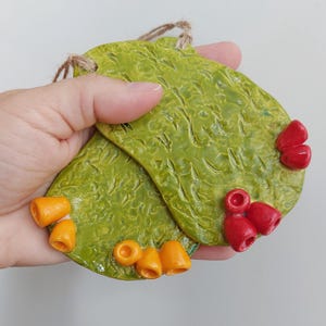 May include: Two flat, oval-shaped ceramic ornaments with a textured, glossy lime green glaze. One ornament features small, bright orange cone-shaped accents, while the other has vibrant red cone-shaped accents. Each piece includes a jute twine loop.