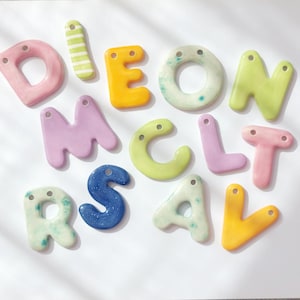 May include: A collection of brightly coloured ceramic letter beads. The letters are in various colours, including pink, yellow, green, blue, and purple. Each letter has a hole for threading. Suitable for jewellery making or crafts.