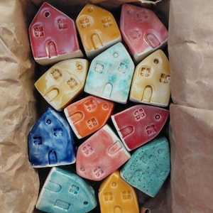 Może przedstawiać: Kolekcja kolorowych ceramicznych miniaturowych domków w brązowym papierowym pojemniku. Domki pomalowane są w różnych kolorach, w tym czerwonym, niebieskim, żółtym i pomarańczowym. Każdy domek ma drzwi i okna.