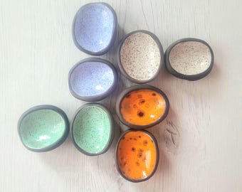 May include: A collection of handmade ceramic bowls in various colors, including blue, green, and orange. Each bowl has a dark rim and a speckled or solid interior. The bowls are arranged on a light-colored wooden surface.