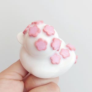 Puede incluir: Una figura de pato de goma blanco decorada con m&uacute;ltiples peque&ntilde;os detalles en relieve en forma de flor de color rosa claro en la cabeza y el cuerpo. El pato tiene un acabado liso y brillante y una forma cl&aacute;sica redondeada.