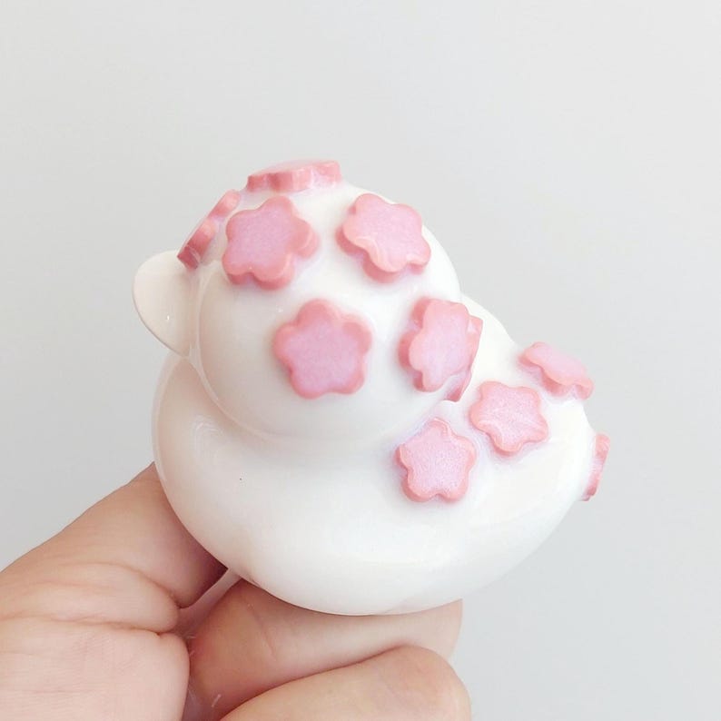 Puede incluir: Una figura de pato de goma blanco decorada con m&uacute;ltiples peque&ntilde;os detalles en relieve en forma de flor de color rosa claro en la cabeza y el cuerpo. El pato tiene un acabado liso y brillante y una forma cl&aacute;sica redondeada.