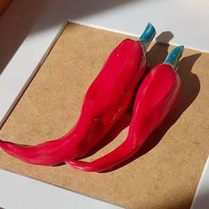 Puede incluir: Dos pimientos rojos brillantes con tallos azul verdoso descansan sobre una superficie marrón claro. Los pimientos están colocados en un ligero ángulo, mostrando su forma curvada. La imagen está enmarcada por un borde blanco.