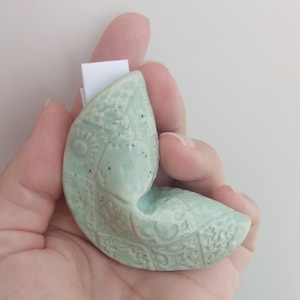 Può includere: Un ornamento in ceramica a forma di biscotto della fortuna color verde menta chiaro, caratterizzato da un motivo in rilievo con forme geometriche e disegni floreali, con un piccolo foglietto di carta bianco inserito nell'apertura.