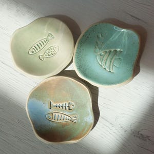 Può includere: Tre piatti in ceramica fatti a mano nei toni del verde, blu e marrone. Ogni piatto presenta un esclusivo design a rilievo di pesce. I piatti sono di forma irregolare e sembrano smaltati, con un'estetica rustica e artigianale.