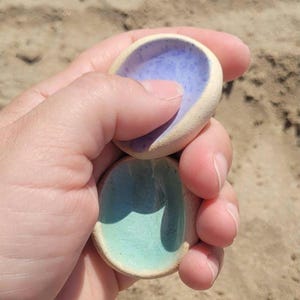 May include: Two small, rounded ceramic dishes with a matte, unglazed beige exterior. One dish features a smooth, glossy interior glaze in a light lavender purple, while the other dish has a glossy interior glaze in a soft, pale mint green color.