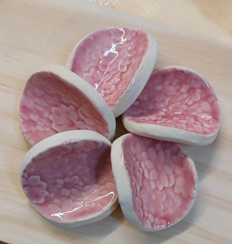 May include: Five small, petal-shaped ceramic dishes. The interior of each dish is a glossy pink, decorated with a floral pattern. The rims are white. These decorative dishes could be used for serving small portions of food or as decorative accents.