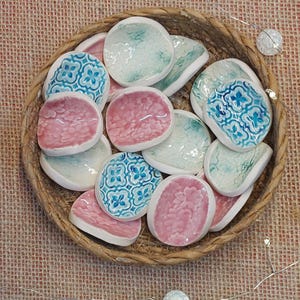 May include: A collection of small, ceramic dishes in a woven basket. The dishes are in shades of pink, teal, and white, with some featuring a floral pattern in blue. The basket is made of natural-colored woven material.