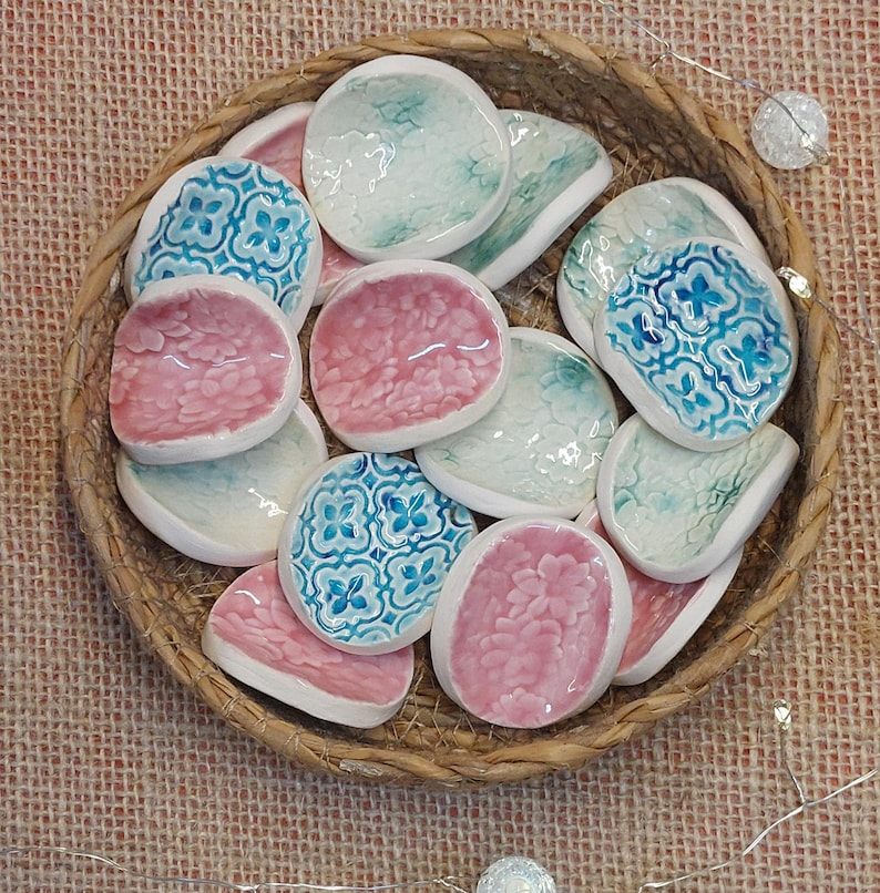 May include: A collection of small, ceramic dishes in a woven basket. The dishes are in shades of pink, teal, and white, with some featuring a floral pattern in blue. The basket is made of natural-colored woven material.