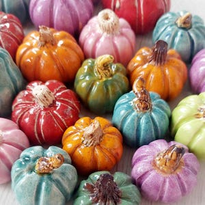 Op de afbeelding: Een verzameling kleurrijke keramische pompoenen in verschillende tinten, waaronder oranje, roze, groen en blauw. Elke pompoen heeft een textuuroppervlak en een bruine steel. De pompoenen zijn dicht bij elkaar geplaatst, waardoor een feestelijke herfsttentoonstelling ontstaat.