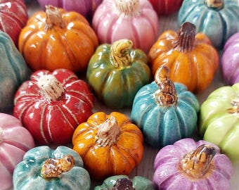 May include: A collection of small, glazed ceramic pumpkins in various colors including orange, red, purple, pink, green, and blue. Each miniature pumpkin features a distinct ribbed texture and a small, textured brown stem on top.
