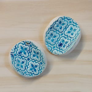 May include: Two ceramic soap dishes with a white base and a blue floral pattern. The dishes have an oval shape and are placed on a light wood surface. The blue pattern is a repeating design of stylized flowers.