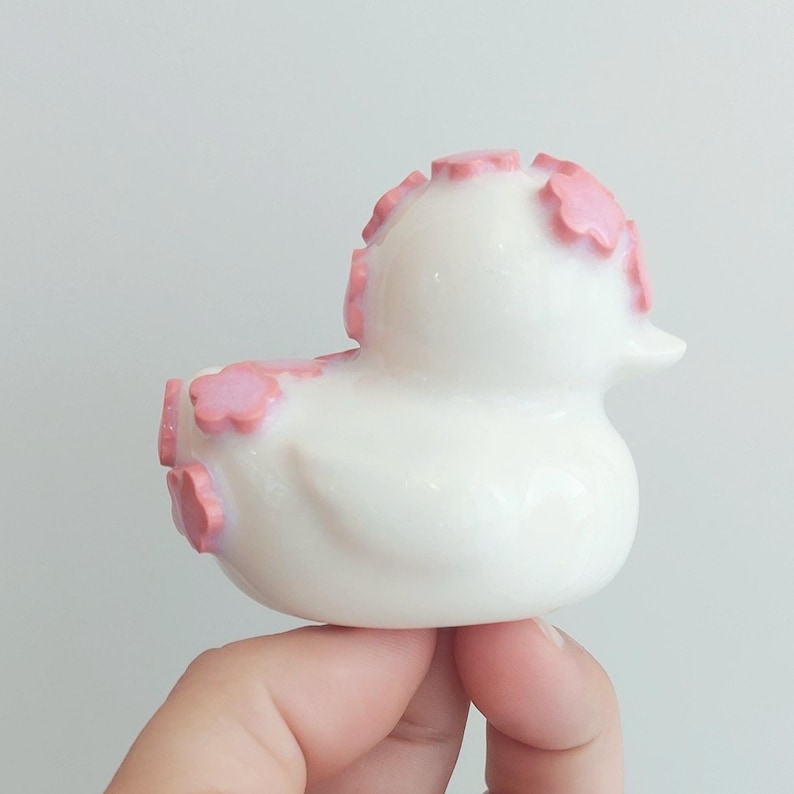 Puede incluir: Una peque&ntilde;a figura de pato de goma blanco con un acabado liso y brillante. El pato est&aacute; decorado con varios detalles en relieve en forma de flor de color rosa claro aplicados en la parte superior de la cabeza, el lomo y la cola.