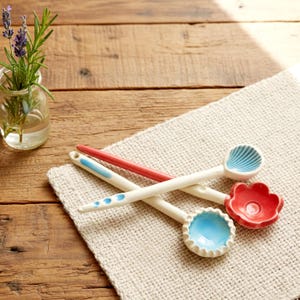 May include: Three decorative ceramic spoons with white handles and colourful bowls. One spoon has a red handle and a flower-shaped bowl, another has a blue bowl with a scalloped edge, and the third has a seashell-shaped bowl.
