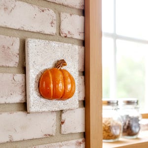 May include: A ceramic pumpkin decoration with an orange glossy finish and a textured brown stem. The pumpkin is mounted on a speckled white square tile, attached to a white brick wall. The image evokes a fall or harvest theme.