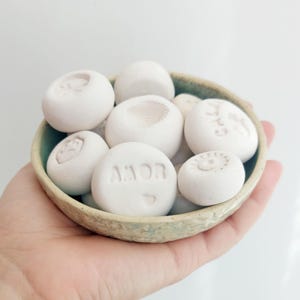 Puede incluir: Un pequeño cuenco de cerámica verde claro lleno de piedras blancas y redondas. Las piedras tienen varios diseños en relieve, incluyendo la palabra "AMOR" y otros patrones. El cuenco se sostiene en una mano sobre un fondo blanco.