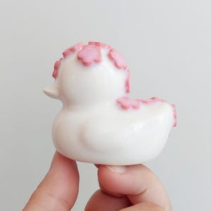 Puede incluir: Una peque&ntilde;a figura de pato de goma blanco con un acabado liso y brillante. El pato est&aacute; decorado con varios detalles en relieve en forma de flor de color rosa claro colocados en la parte superior de la cabeza y a lo largo del lomo.