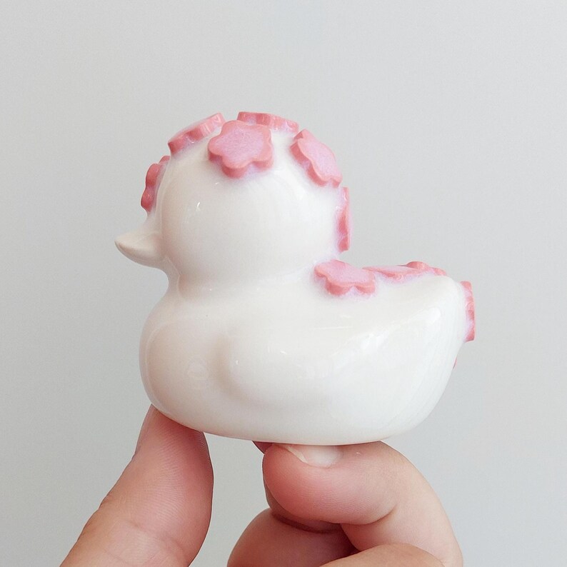 Puede incluir: Una peque&ntilde;a figura de pato de goma blanco con un acabado liso y brillante. El pato est&aacute; decorado con varios detalles en relieve en forma de flor de color rosa claro colocados en la parte superior de la cabeza y a lo largo del lomo.