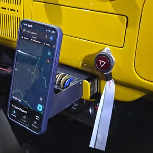 Può includere: Uno smartphone blu in una custodia mostra una mappa, montato su un cruscotto giallo. Un portamonete è sotto il telefono. Un pulsante rosso e argento con un triangolo bianco è a destra.