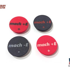 May include: Four circular objects with the text "mach +E", "mach -E", and a plus or minus symbol. Two are red, two are black. The objects are on a white surface. The image also includes the text "ASBURY SOLUTION" and "MADE IN USA".