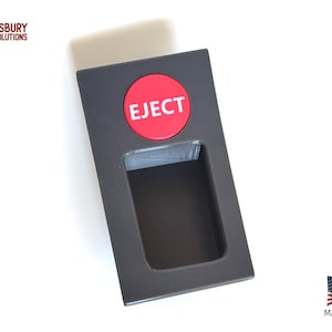 May include: A rectangular, dark gray plastic component with a red circular button labeled "EJECT". The component has a rectangular opening and is set against a white background. The text "ABS Asbury Solutions" is in the upper left corner.